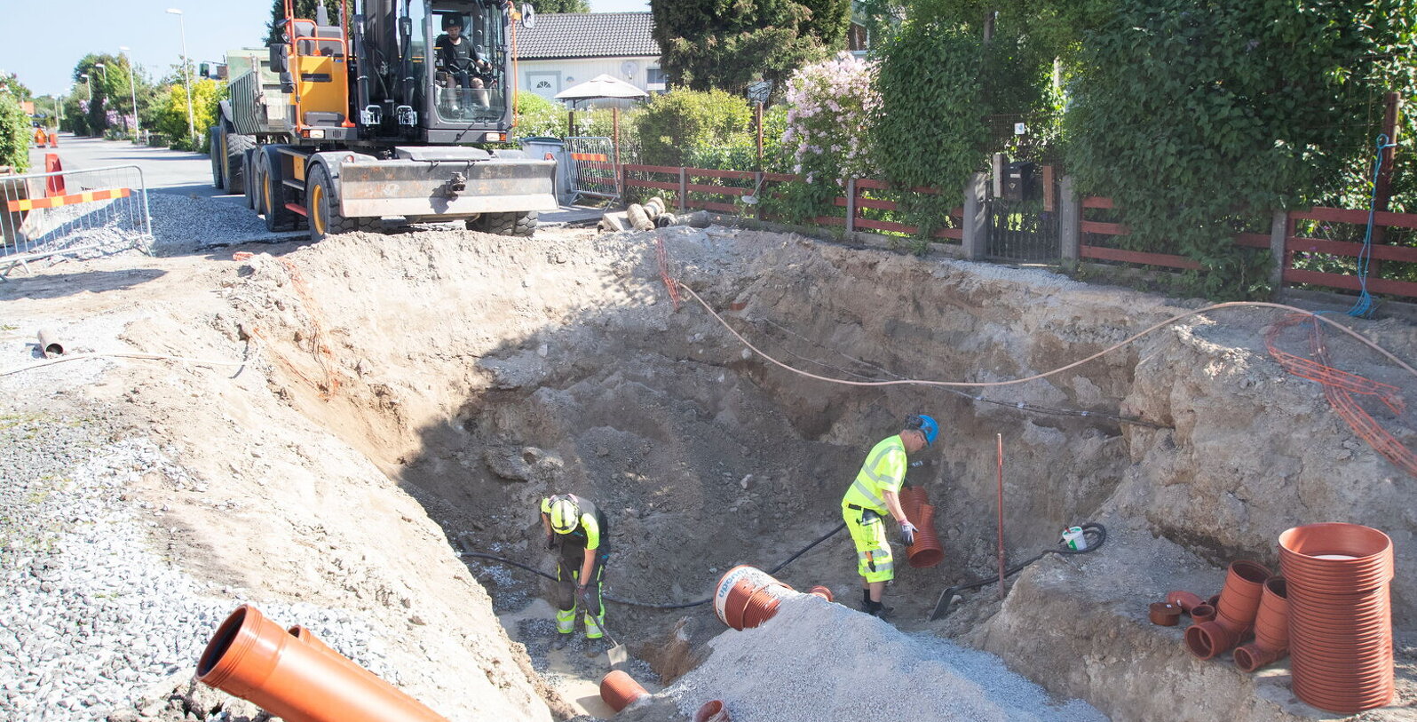 Bild på en grävmaskin och två medarbetare på SBVT som installerar nya rör under en uppgrävd väg.